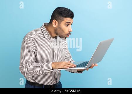 Porträt eines schockierten Geschäftsmannes mit Bart, der auf ein Notebook-Display blickt, mit offenem Mund und großen Augen, Systemfehler, gestreiftem Hemd. Innenstudio, isoliert auf blauem Hintergrund. Stockfoto