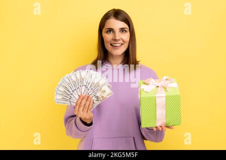 Glückliche optimistische Frau, die eingepackte Geschenkschachtel und Dollarscheine in der Hand hält, mit positivem Ausdruck in die Kamera schaut, lila Kapuzenpulli trägt. Innenstudio-Aufnahme isoliert auf gelbem Hintergrund. Stockfoto