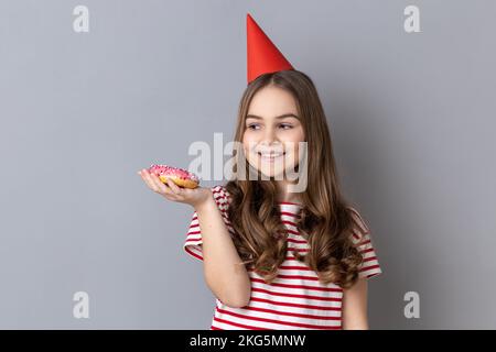 Porträt eines fröhlichen kleinen Mädchens, das gestreiftes T-Shirt und Partykegel trägt, auf der Hand hält, leckeren süßen Donut hält und beim Dessert lächelt. Studioaufnahmen im Innenbereich isoliert auf grauem Hintergrund. Stockfoto