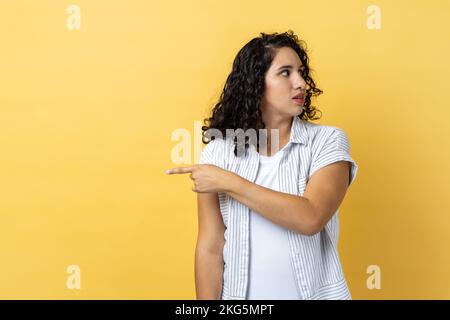 Porträt einer Frau mit dunklem, wellenförmigem Haar, die mit dem Finger zur Seite zeigt, die Ausstieg angeordnet hat und verärgert aussieht, der Boss von der Arbeit abtritt, den Ausstieg zeigt. Innenstudio-Aufnahme isoliert auf gelbem Hintergrund. Stockfoto