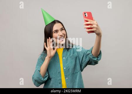 Porträt einer attraktiven Frau mit Partykegel, die Selfie macht oder Livestream überträgt, mit der Hand winkt, hallo sagt, eine lässige Jacke trägt. Studioaufnahmen im Innenbereich isoliert auf grauem Hintergrund. Stockfoto