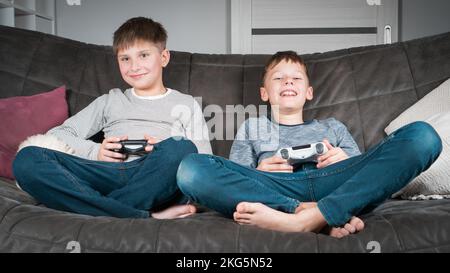 Porträt von zwei glücklichen, zufriedenen Jungen im Teenageralter, die zu Hause auf dem Sofa sitzen, den Joystick des Gaming-Controllers halten, Videospiele spielen, Spaß haben. Hobby, frei Stockfoto