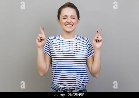 Portrait einer hoffnungsvollen, schönen, jungen erwachsenen Frau, die gestreiftes T-Shirt trägt und die Finger kreuzt, Hoffnung auf Besseres, Glückwunsch, Ritual. Studioaufnahmen im Innenbereich isoliert auf grauem Hintergrund. Stockfoto