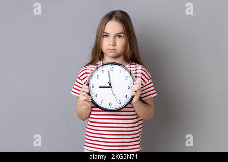 Porträt eines ernsten kleinen Mädchens, das gestreiftes T-Shirt trägt, eine große Wanduhr hält, mit unangenehmen Gefühlen in die Kamera schaut, Zeit zu gehen. Studioaufnahmen im Innenbereich isoliert auf grauem Hintergrund. Stockfoto