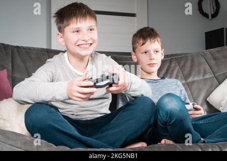 Porträt von zwei lächelnden, glücklichen Jungen im Teenageralter, die zu Hause auf einem grauen Sofa sitzen, den Joystick des Gaming-Controllers halten, Videospiele spielen und Spaß haben. Hobby, frei Stockfoto