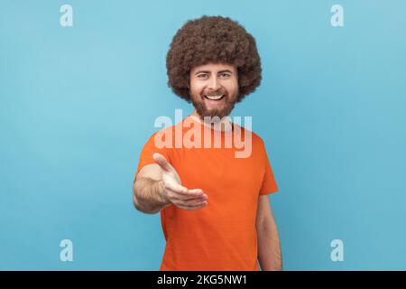 Porträt eines Mannes mit Afro-Frisur, der ein orangefarbenes T-Shirt trägt, mit ausgestreckter Hand steht, dem Partner einen Handschlag gibt, während des Vorstellungsgesprächs begrüßt wird. Innenstudio, isoliert auf blauem Hintergrund. Stockfoto