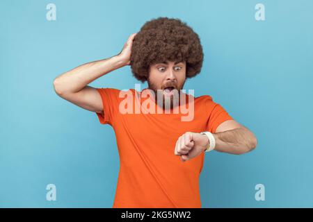 Porträt eines Mannes mit Afro-Frisur, der ein orangefarbenes T-Shirt trägt und mit dem Finger auf seine Armbanduhr zeigt, mit nervösem Gesicht, die Zeit ist abgelaufen, hält die Hand am Kopf. Innenstudio, isoliert auf blauem Hintergrund. Stockfoto