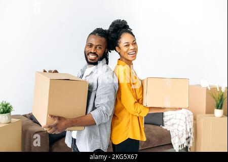 Glückliches Paar aus afroamerikanischer Familie, das Kisten mit Sachen für das Haus hält, mit dem Rücken zueinander steht, in die Kamera schaut, lächelt, sich freut, eine eigene Wohnung oder ein Haus zu kaufen Stockfoto