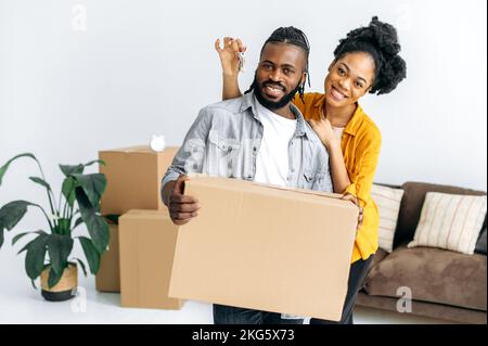 Glücklicher Mann und Frau, stehen in ihrem neuen Wohnzimmer, Mann mit großer Schachtel, Frau mit Schlüssel, sie schauen in die Kamera, lächeln. Ein liebenswertes afroamerikanisches Ehepaar zieht in sein neues Zuhause Stockfoto