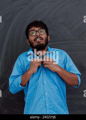 Ein indischer junger Schüler mit Brille zieht ein blaues Hemd an, während er vor der Schultafel steht Stockfoto