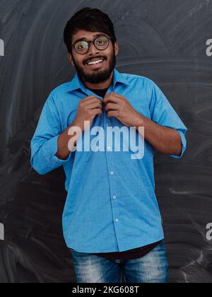 Ein indischer junger Schüler mit Brille zieht ein blaues Hemd an, während er vor der Schultafel steht Stockfoto