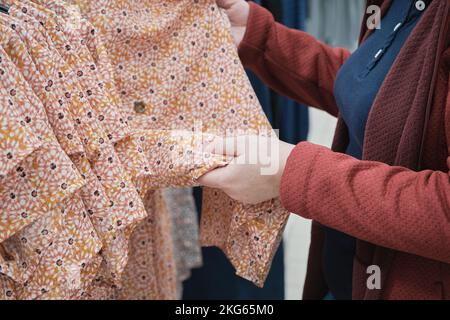 Eine schwangere Frau sucht sich ein lockeres Kleid in einer Blume zum Kauf aus. Hände aus der Nähe Stockfoto