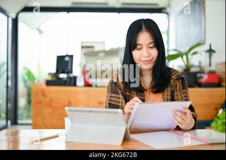 Charmante junge asiatische Freiberuflerin für College-Studentin, die im Café arbeitet und etwas über Papierkram analysiert. Stockfoto