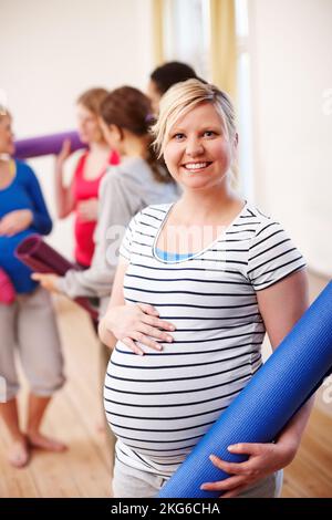 Während der Schwangerschaft in Form bleiben. Eine junge blonde Schwangere lächelt, während sie ihre Trainingsmatte hält, während ihre Freunde im Hintergrund stehen. Stockfoto