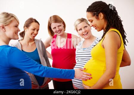 Wir können es kaum erwarten, den Kleinen zu treffen. Eine junge Frau berührt den Bauch ihrer Schwangeren, während sie in einer Gruppe steht und eine freundliche Diskussion führt. Stockfoto