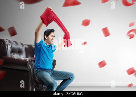 Asiatischer Mann mit einem Schal, der auf der Couch sitzt und mit einem aufgeregten Ausdruck zu Hause Fußball im Fernsehen guckt Stockfoto