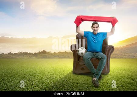 Ein asiatischer Mann mit dem Ball, der den Schal hält, sitzt auf der Couch und schaut sich Fußball im Freien an Stockfoto