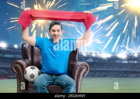 Asiatischer Mann mit dem Ball hält Schal sitzt auf der Couch mit einem aufgeregt Ausdruck im Fußballstadion Hintergrund Stockfoto