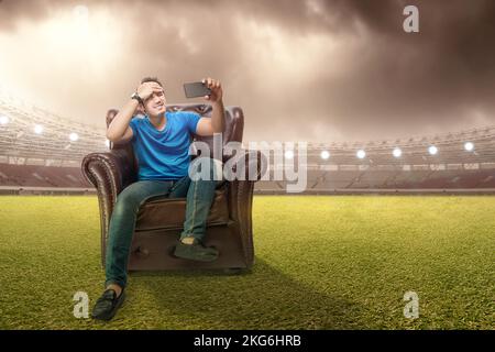 Asiatischer Mann mit dem Ball sitzt auf der Couch mit einem enttäuschten Ausdruck beobachten ein Fußballspiel von einem Handy im Fußballstadion zurück Stockfoto