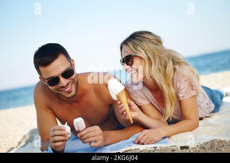 Esst es, bevor es schmilzt. Ein glückliches junges Paar, das Eis isst, während es an einem sonnigen Strand liegt. Stockfoto