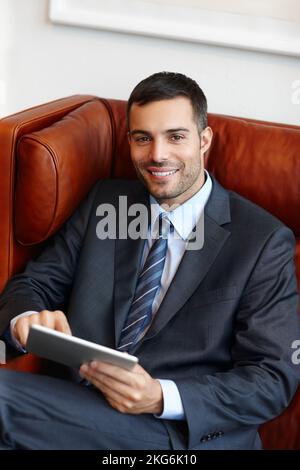 Mit modernen Werkzeugen, um seinen Erfolg zu unterstützen. Ein lächelnder junger Geschäftsmann, der auf einem Ledersofa sitzt, während er ein digitales Tablet benutzt. Stockfoto
