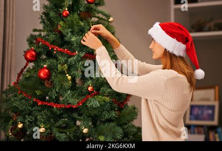 Die letzten Berührungen auf den Baum setzen. Eine schöne junge Frau, die ihren Weihnachtsbaum zu Hause schmückt. Stockfoto