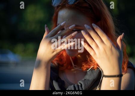 Das Mädchen hält ihre Jungs vor ihr Gesicht. Das Mädchen versteckt sich hinter ihren Händen und lacht. Schüchterne Persönlichkeit. Lange Nägel. Stockfoto