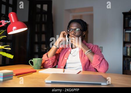 Gestresster afroamerikanischer Freiberufler, der telefoniert, sitzt mit Laptop und Tasse am Tisch Stockfoto