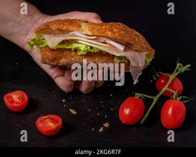 Eine weibliche Hand hält ein Sandwich gefüllt mit Käse, Salat und geräuchertem Schinken auf schwarzem Hintergrund mit roten Kirschtomaten Stockfoto