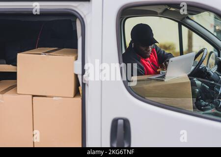 Außenansicht eines fokussierten, hart arbeitenden 30-jährigen afroamerikanischen Mannes, der an seinem Laptop im Paketwagen arbeitet. Seitentür des weißen Lieferwagens mit vielen Kartonpaketen, die zur Auslieferung bereit sind. Hochwertiges Foto Stockfoto