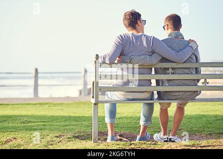 Liebe am Meer. Rückansicht eines jungen schwulen Paares, das zusammen auf einer Parkbank sitzt. Stockfoto