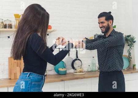 Ein junges, fröhliches, elegantes Paar, das lacht, Händchen hält, in der Küche in seiner neuen hellen Wohnung tanzt, den Umzug feiert. Hochwertiges Foto Stockfoto