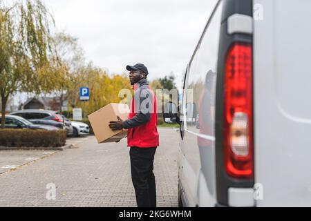 Gut gebauter professioneller afroamerikanischer männlicher Kurier mit einem Pappkarton, der nach der richtigen Adresse sucht, steht vor seinem weißen Lieferwagen. Outdoor-Aufnahmen. Anfang Herbst. Hochwertiges Foto Stockfoto