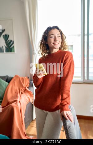Lächelnde junge Frau, die ihr Handy benutzt und zu Hause im Wohnzimmer steht. Stockfoto