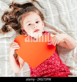 Porträt eines lächelnden kleinen Mädchens, das auf einem hellen Bettüberwurf liegt und eine Valentinskarte mit großem Herz in der Hand hält. Draufsicht Stockfoto