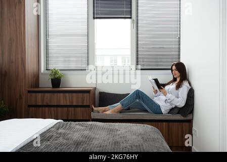 Frau liest Buch in einem Schlafzimmer mit Bett und Holzgarderobe in einem modernen Apartment Stockfoto