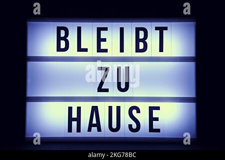 Ein Leuchtkasten mit der Meldung „ZU HAUSE BLEIBEN“ in deutschen schwarzen Buchstaben, beleuchtet vor dunklem Hintergrund. Die Nachricht ist im Mittelpunkt des Bildes. Stockfoto