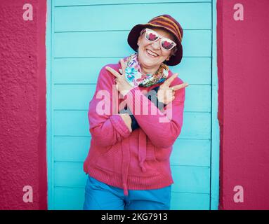 Porträt, Ruhe und Funky mit einer älteren Frau im Freien, die vor einer blauen Tür und einem roten Wandhintergrund steht. Brille, Hände und Hip Hop mit einem Happy Stockfoto