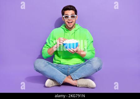 Foto in voller Länge von einem beeindruckenden, witzigen Kerl in Neon-Sweatshirt, der Popcorn isst und einen Film mit einem isolierten violetten Hintergrund ansieht Stockfoto