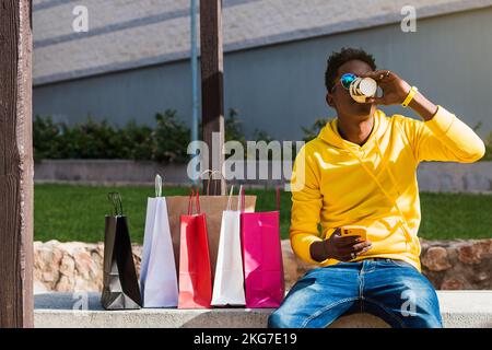 Junger afrikanischer Mann, der auf einer Parkbank sitzt und Kaffee aus einem Pappbecher trinkt, während er sein Handy in der Hand hält. Neben ihm sind mehrere Papiereinkäufe Stockfoto