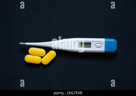 Elektronisches Thermometer liegt neben gelben Pillen auf einem isolierten Hintergrund, Medizin und Apotheke, Gesundheit und Behandlung, Pillen und Antibiotika Stockfoto