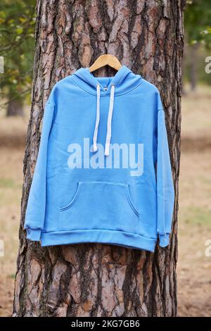 Blauer Kapuzenpullover, der an einem Bügel an einem Baum im Wald hängt. Darstellung verschiedener Farben der Kleidung Stockfoto