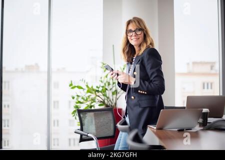 Eine lächelnde Geschäftsfrau mittleren Alters, die während eines Videoanrufs im Büro Smartphone und Kopfhörer verwendet. Stockfoto
