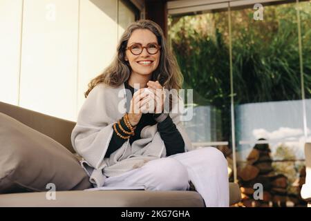 Entspannen Sie sich bei einer heißen Tasse Tee. Reife Frau, die fröhlich lächelt, während sie Tee auf einer Couch trinkt. Glückliche Seniorin, die einen ruhigen Morgen in ihrer Liv genießt Stockfoto