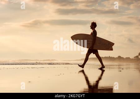 Ein hübscher Surfer geht mit Surfbrett in den Händen zum Strand bei Sonnenuntergang Stockfoto