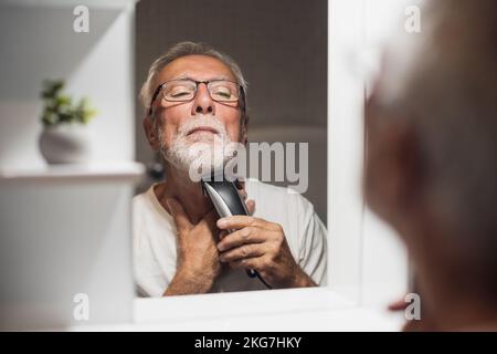 Der Seniorist schneidet seinen Bart mit einem elektrischen Trimmer im Badezimmer. Stockfoto