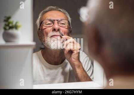 Der Seniorenmann pflückt mit einer Pinzette im Bad laute Haare. Stockfoto