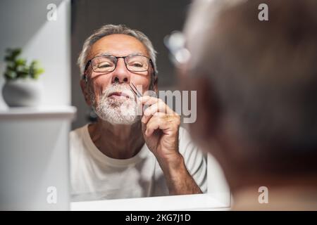 Der Seniorenmann pflückt mit einer Pinzette im Bad laute Haare. Stockfoto