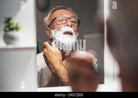 Der Seniorist rasiert sich im Badezimmer mit Rasiermesser und Schaum. Stockfoto