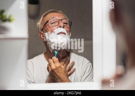 Der Seniorist rasiert sich im Badezimmer mit Rasiermesser und Schaum. Stockfoto